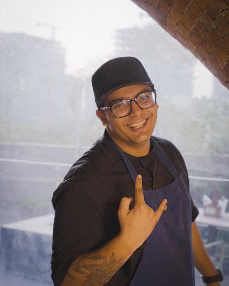 Chef Shlok Shah smiling in a black cap, glasses and a navy apron, standing by a window
